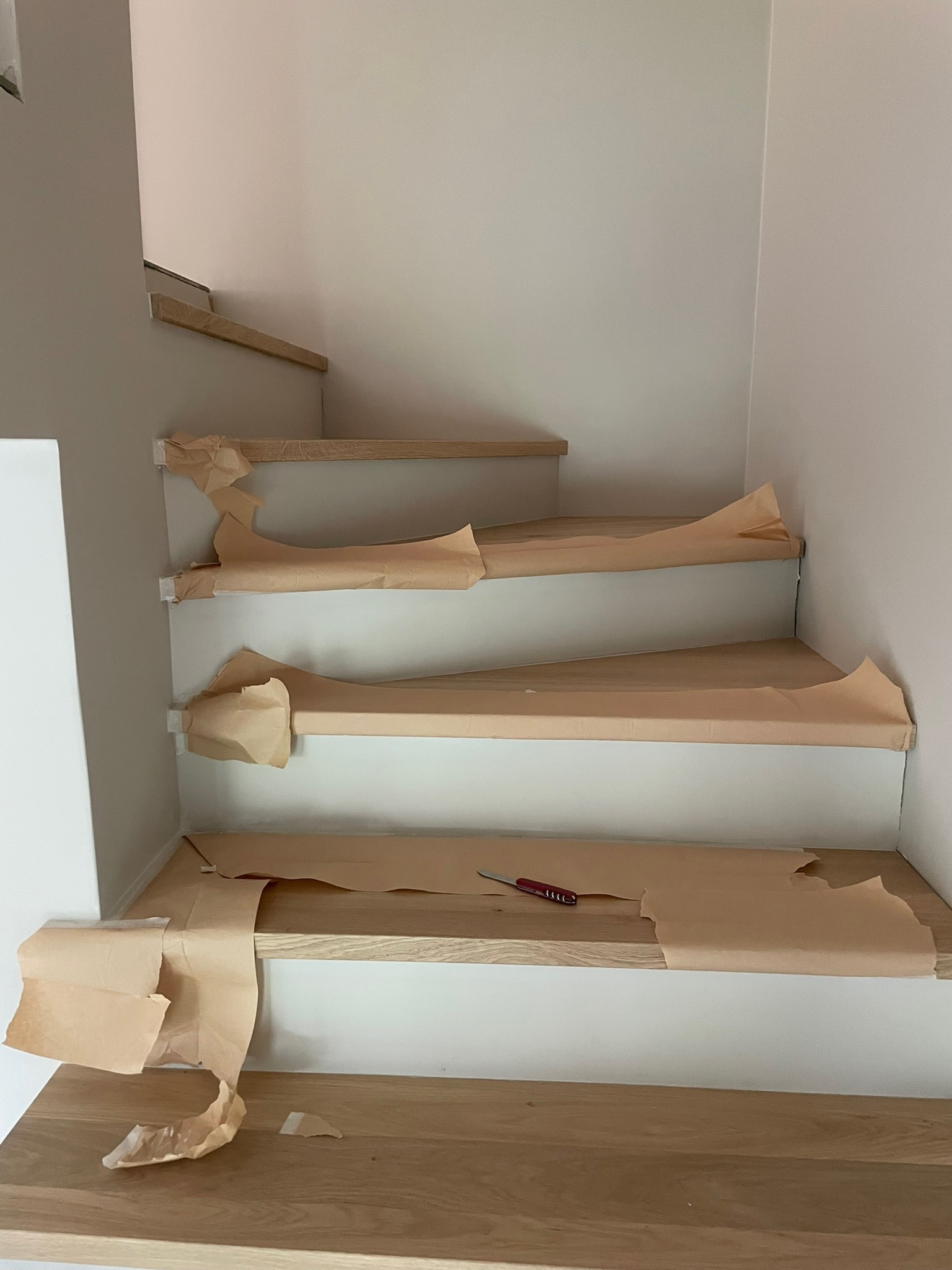Jasne drewniane schody zabezpieczone papierem ochronnym podczas prac wykończeniowych. Na jednym ze stopni leży nóż do tapet.