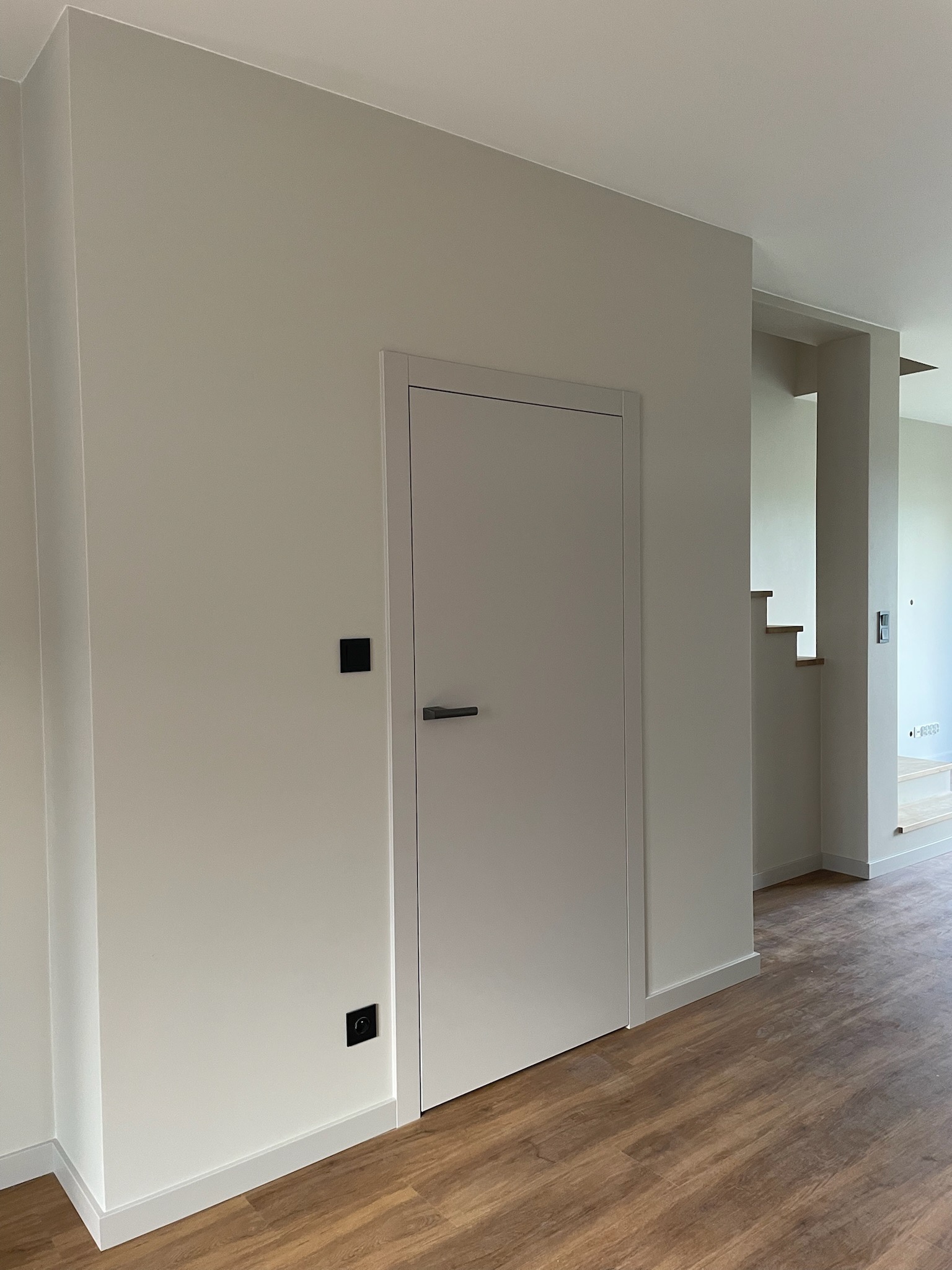 Jasne wnętrze po remoncie: białe ściany, drewnopodobna podłoga, minimalistyczne drzwi z czarną klamką, widoczne schody w tle. Nowoczesne wykończenie.