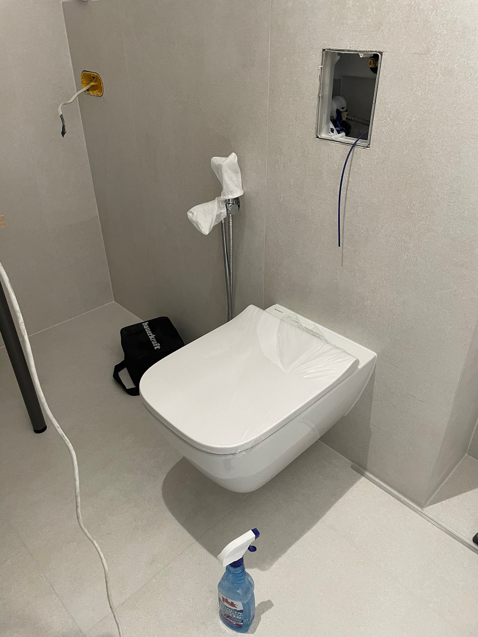 Montaż podwieszanej toalety w łazience z szarą płytką. Widoczne elementy instalacji, narzędzia i zabezpieczenia. Etap prac wykończeniowych.