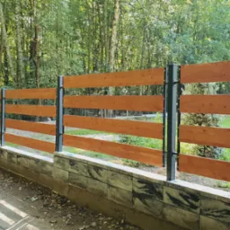 Nowoczesne ogrodzenie drewniane z poziomych desek w ciepłym odcieniu brązu, zamocowanych na metalowych słupkach, osadzone na niskim murku z betonowych bloczków, otoczone zielenią drzew.