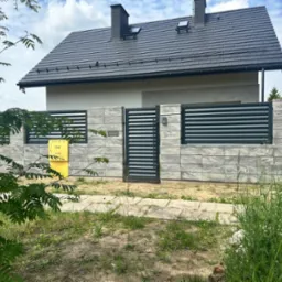 Nowoczesne ogrodzenie panelowe z szarej kostki i metalowych przęseł, brama wejściowa, skrzynka gazowa na tle domu z ciemną dachówką.