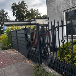 fixed Łukasz Banaś - Nowoczesna, grafitowa brama wjazdowa i ozdobne, metalowe ogrodzenie posesji z widoczną skrzynką na listy, na tle budynku z szarą elewacją i zielenią.