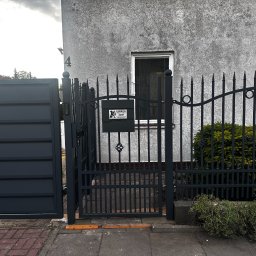 fixed Łukasz Banaś - Ciemnoszara, metalowa brama wjazdowa i furtka z ostrzami, z tabliczką ostrzegawczą 'Uwaga! Teren pilnowany'. W tle szary budynek z oknem i numerem 4.