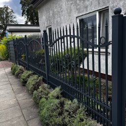 fixed Łukasz Banaś - Eleganckie, grafitowe ogrodzenie metalowe z ozdobnymi elementami, oddzielające posesję od chodnika. W tle budynek z oknem i zielenią.