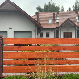 fixed Łukasz Banaś - Nowoczesne ogrodzenie drewniane z ceglanymi słupkami na tle domu z czerwoną dachówką. Widoczne detale drewna i cegły, podkreślone naturalnym światłem.