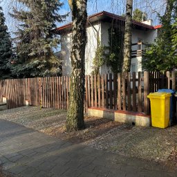fixed Łukasz Banaś - Drewniane ogrodzenie posesji z furtką, widoczne z chodnika. Obok mur z cegły i pojemniki na śmieci. W tle dom z balkonem i drzewa.