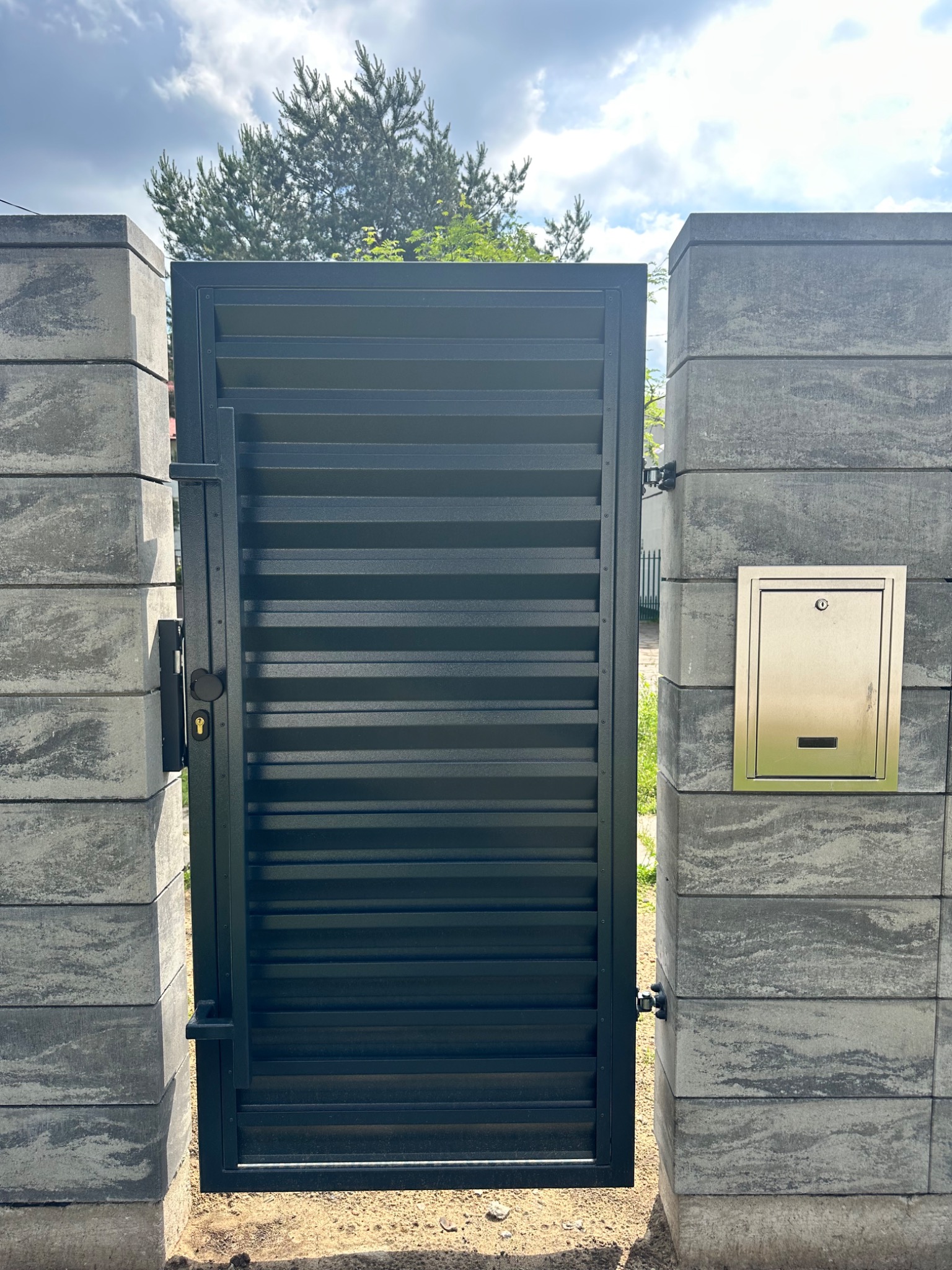 Nowoczesna, czarna furtka z poziomymi lamelami, wkomponowana w szare, betonowe słupki z zamontowaną skrzynką na listy ze stali nierdzewnej.