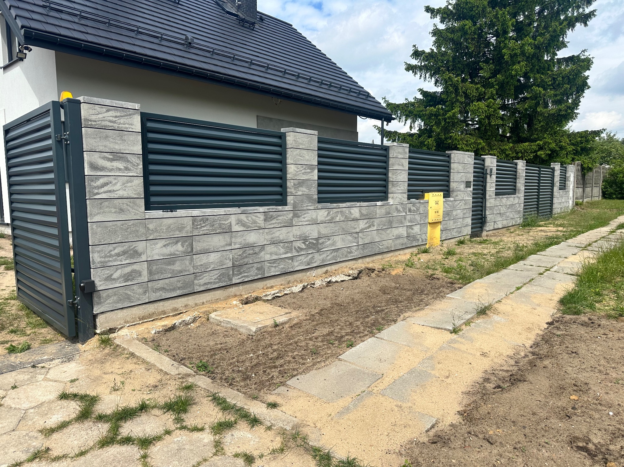 Nowoczesne ogrodzenie panelowe z betonowych bloczków i metalowych przęseł, brama wjazdowa z żółtą lampą ostrzegawczą, widok na dom i skrzynkę gazową.