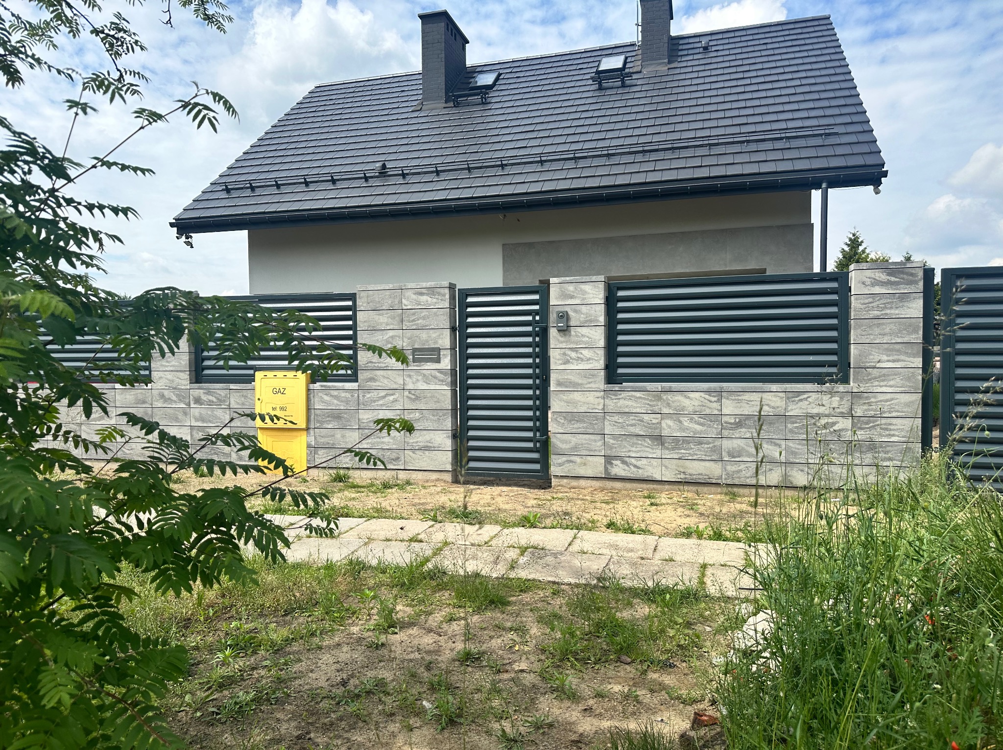Nowoczesne ogrodzenie panelowe z szarej kostki i metalowych przęseł, brama wejściowa, skrzynka gazowa na tle domu z ciemną dachówką.