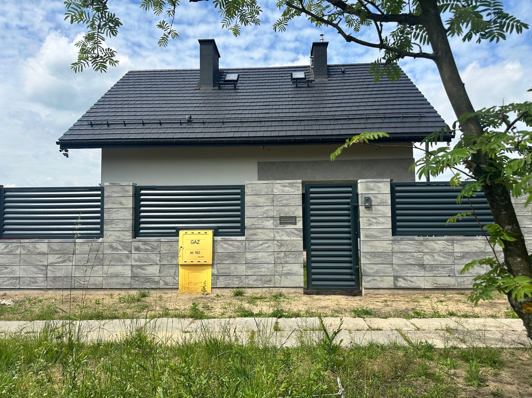 Nowoczesne ogrodzenie panelowe z szarej kostki, zintegrowane z bramką. Żółta skrzynka gazowa przy ogrodzeniu. Dom z ciemną dachówką w tle, częściowo zasłonięty przez drzewo.