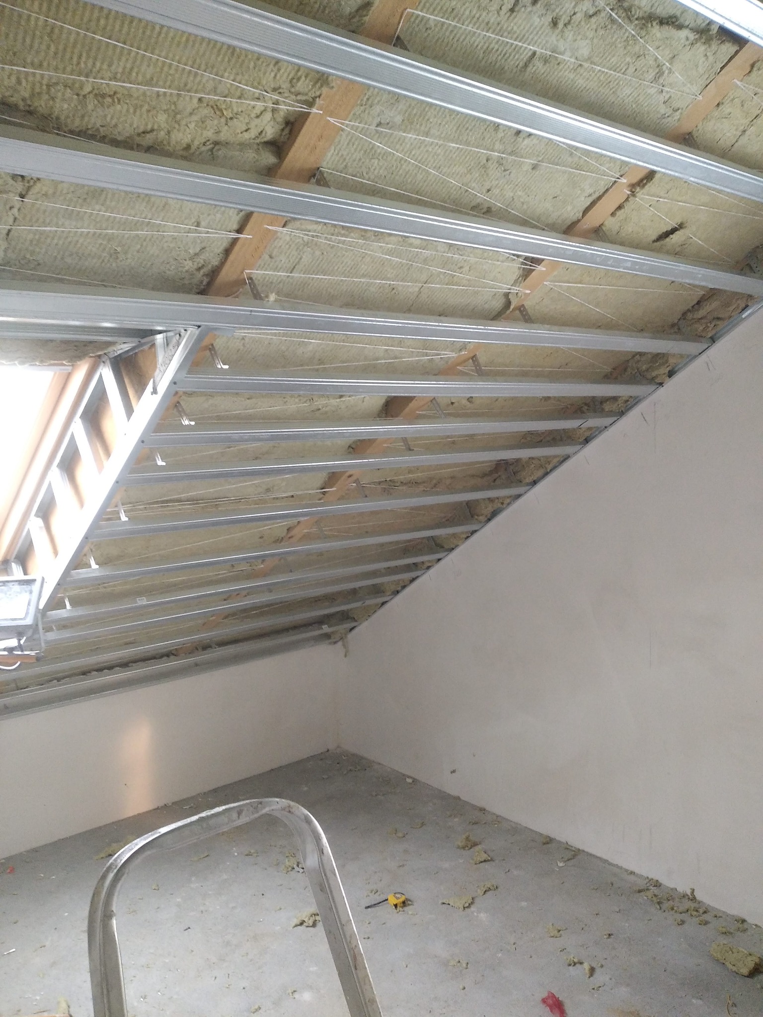 Montaż izolacji z wełny mineralnej na poddaszu, widoczne metalowe profile konstrukcyjne, drewniane elementy więźby dachowej, białe sznurki podtrzymujące izolację oraz fragment ściany i drabiny.