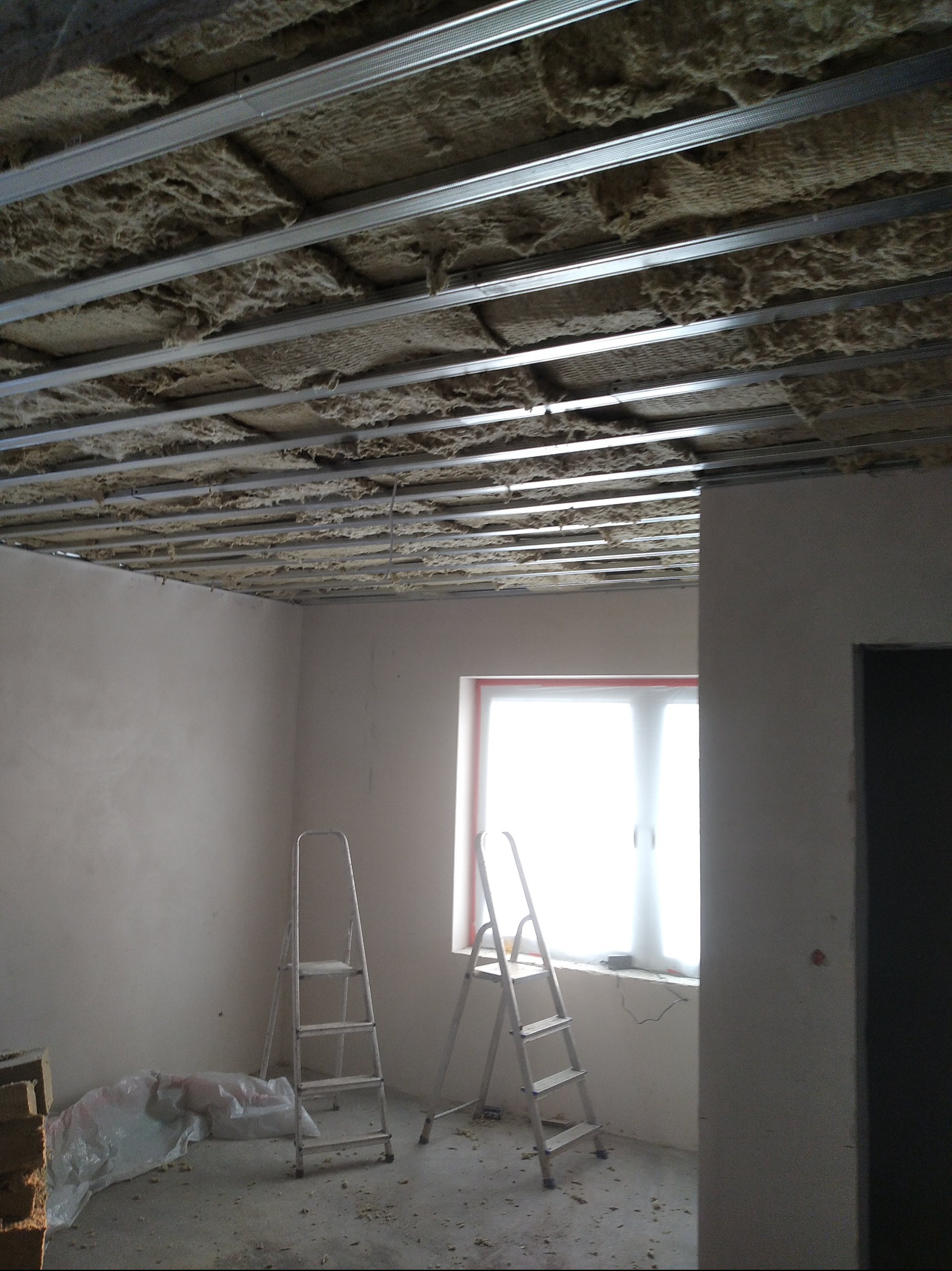 Montaż izolacji z wełny mineralnej pod sufitem podwieszanym, widoczne metalowe profile, dwie aluminiowe drabiny, okno z białymi zasłonami i fragment ściany w surowym stanie.