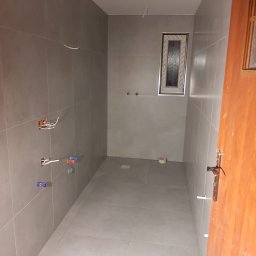 mgr. inż. Renata Jankowska Renovations Company - Wnętrze łazienki w trakcie prac wykończeniowych, widoczne szare płytki na ścianach i podłodze, przygotowane instalacje wodne i elektryczne, oraz małe okno z białymi ramami. Częściowo widoczne...