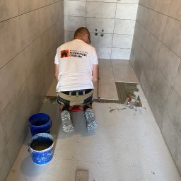 mgr. inż. Renata Jankowska Renovations Company - Pracownik w koszulce firmowej klęczy na czerwonym stołku, układając szare płytki podłogowe w surowej łazience, z widocznymi niebieskimi wiaderkami i narzędziami.