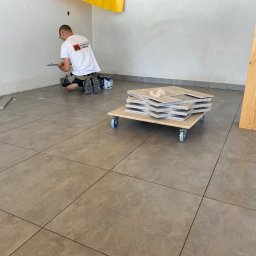 mgr. inż. Renata Jankowska Renovations Company - Pracownik w koszulce z logo firmy układa szare płytki podłogowe w pomieszczeniu, na podłodze leżą płytki na wózku na kółkach, widoczne narzędzia i puszki z farbą.