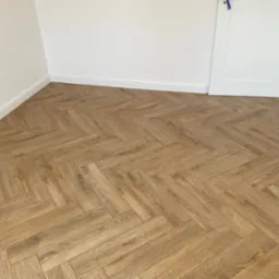 Ułożona pod kątem 45 stopni parkietowa podłoga w odcieniach brązu, z widoczną fakturą drewna, w jasnym pomieszczeniu z białymi ścianami i listwami przypodłogowymi, oraz białe drzwi z niebieską...