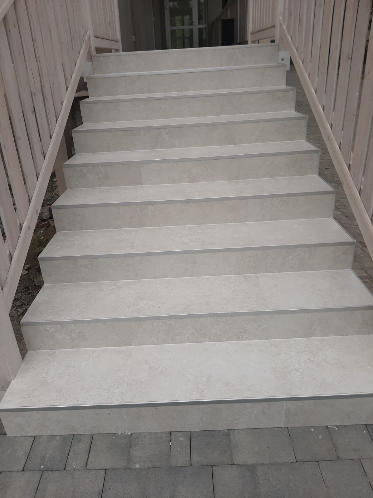 Schody zewnętrzne wyłożone jasnymi płytkami ceramicznymi z metalowymi listwami zabezpieczającymi krawędzie, drewniana balustrada po bokach.