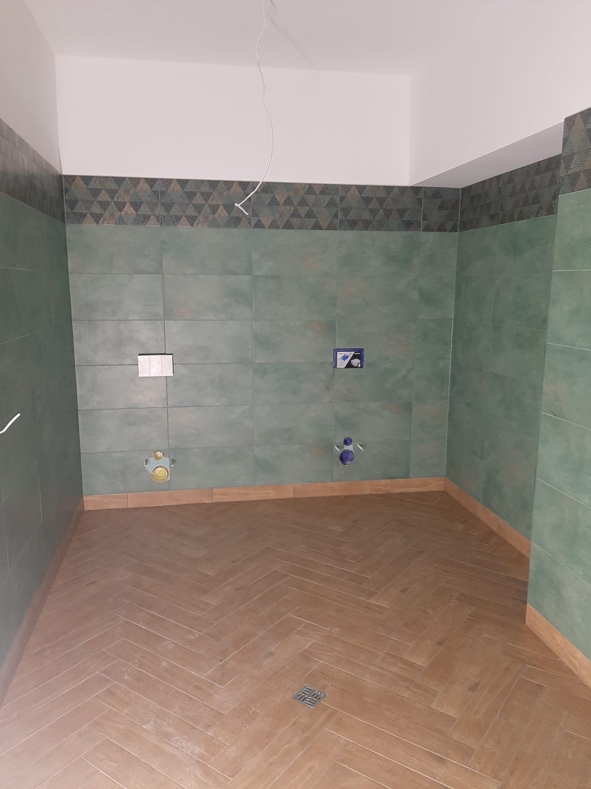 Wnętrze łazienki w Głuchołazach po remoncie, z zielonymi płytkami na ścianach i podłogą wyłożoną parkietem w jodełkę. Widoczne przygotowane przyłącza wodno-kanalizacyjne.