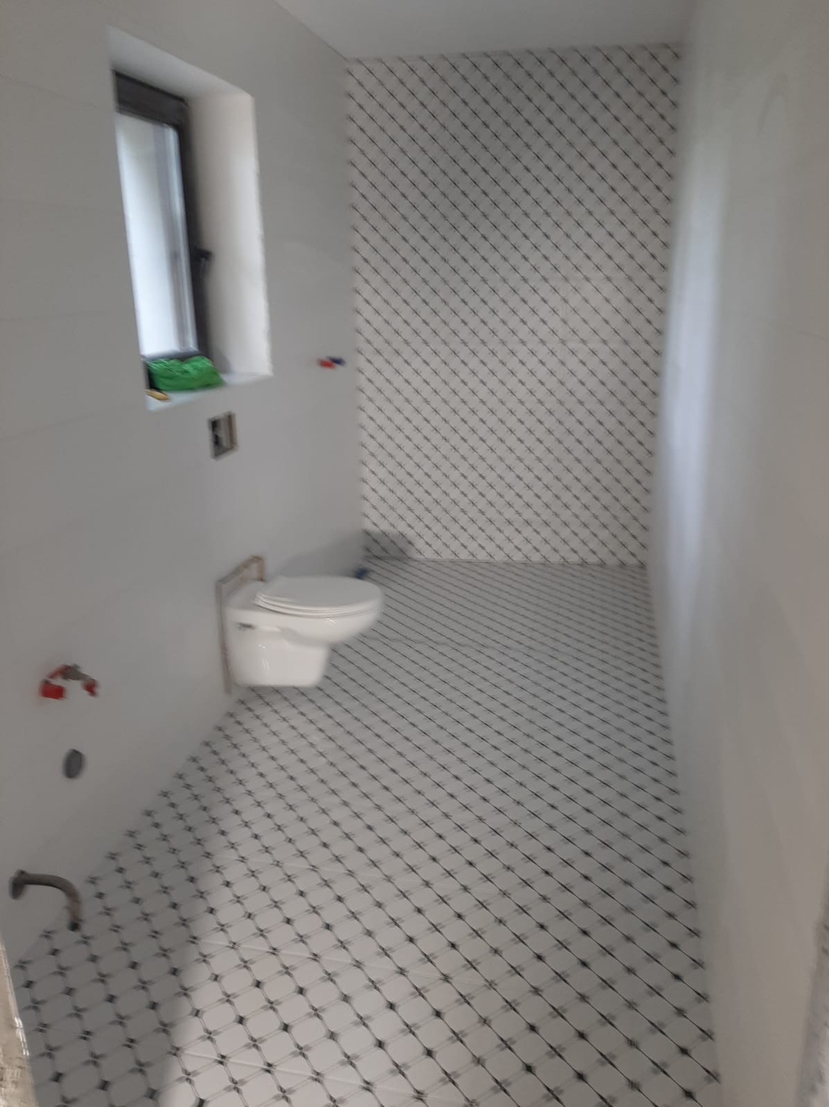 Wnętrze łazienki po remoncie, z białymi płytkami ułożonymi w romby z czarnymi akcentami na podłodze i ścianie, wisząca toaleta oraz okno z zieloną gąbką na parapecie.