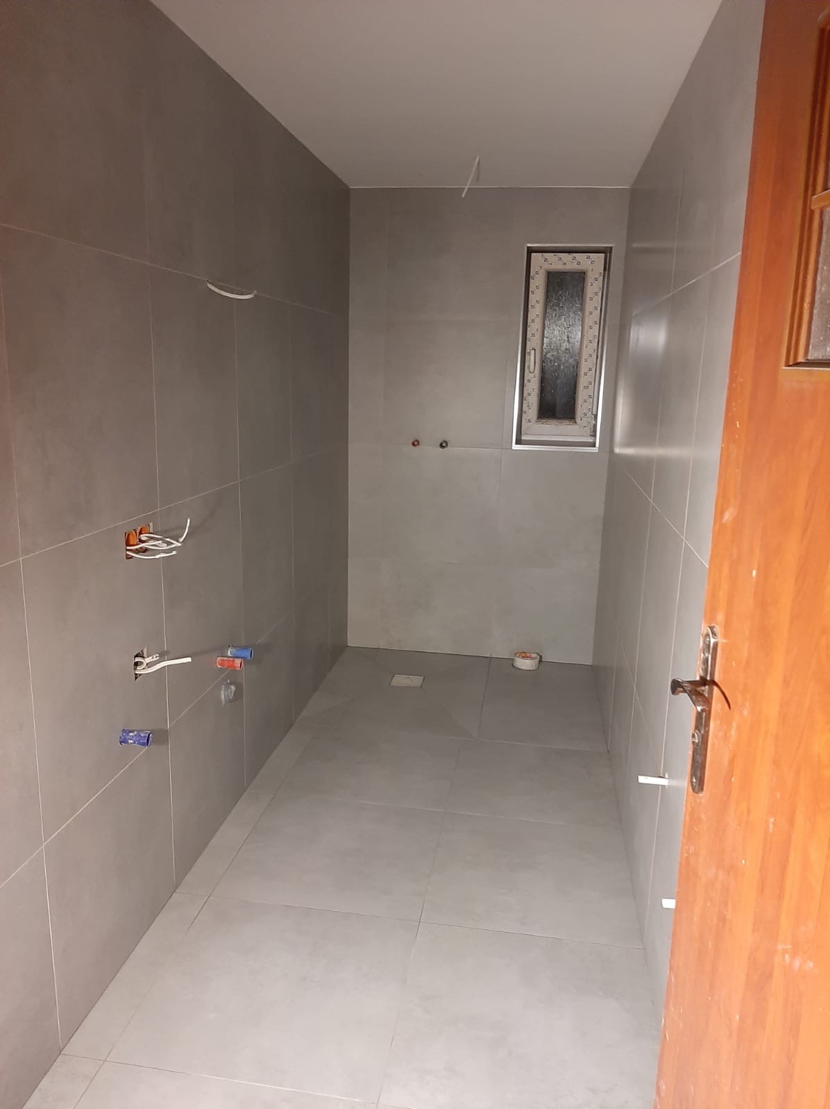 Wnętrze łazienki w trakcie prac wykończeniowych, widoczne szare płytki na ścianach i podłodze, przygotowane instalacje wodne i elektryczne, oraz małe okno z białymi ramami. Częściowo widoczne...