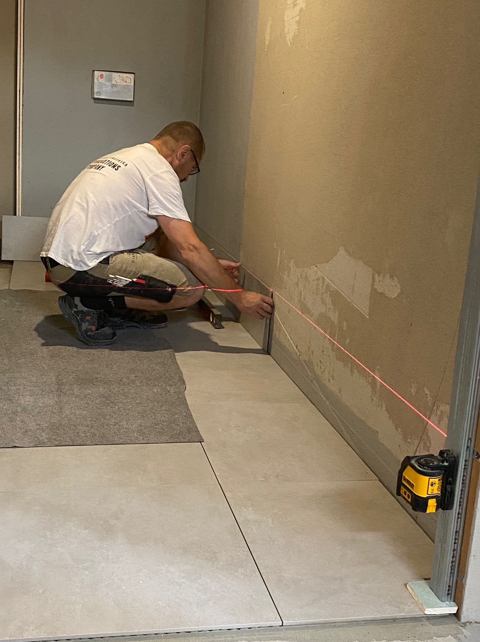 Pracownik w trakcie układania płytek przypodłogowych, używając poziomicy laserowej do wyznaczenia linii, widoczne niedokończone ściany i ułożona podłoga z płytek.