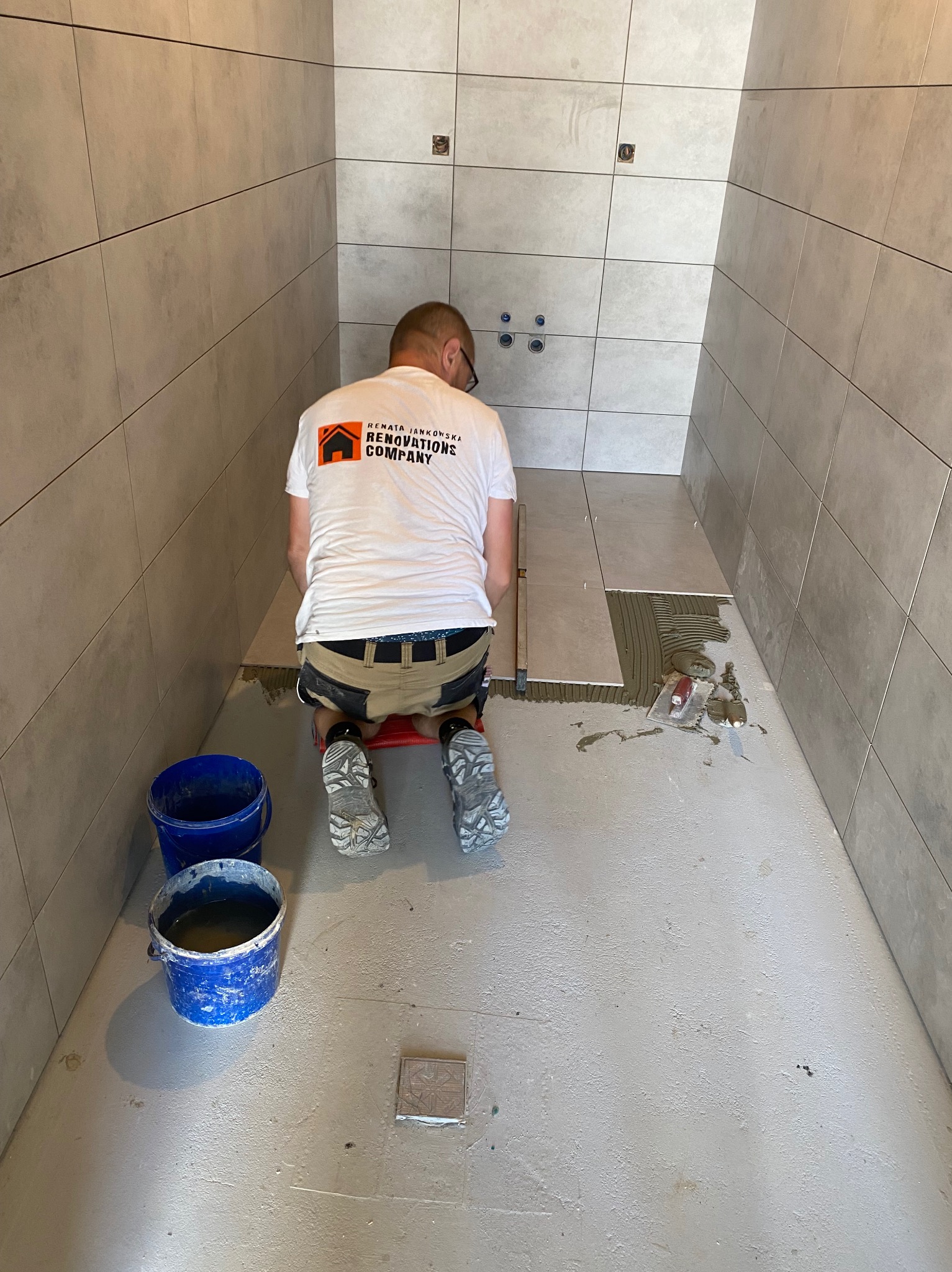 Pracownik w koszulce firmowej klęczy na czerwonym stołku, układając szare płytki podłogowe w surowej łazience, z widocznymi niebieskimi wiaderkami i narzędziami.