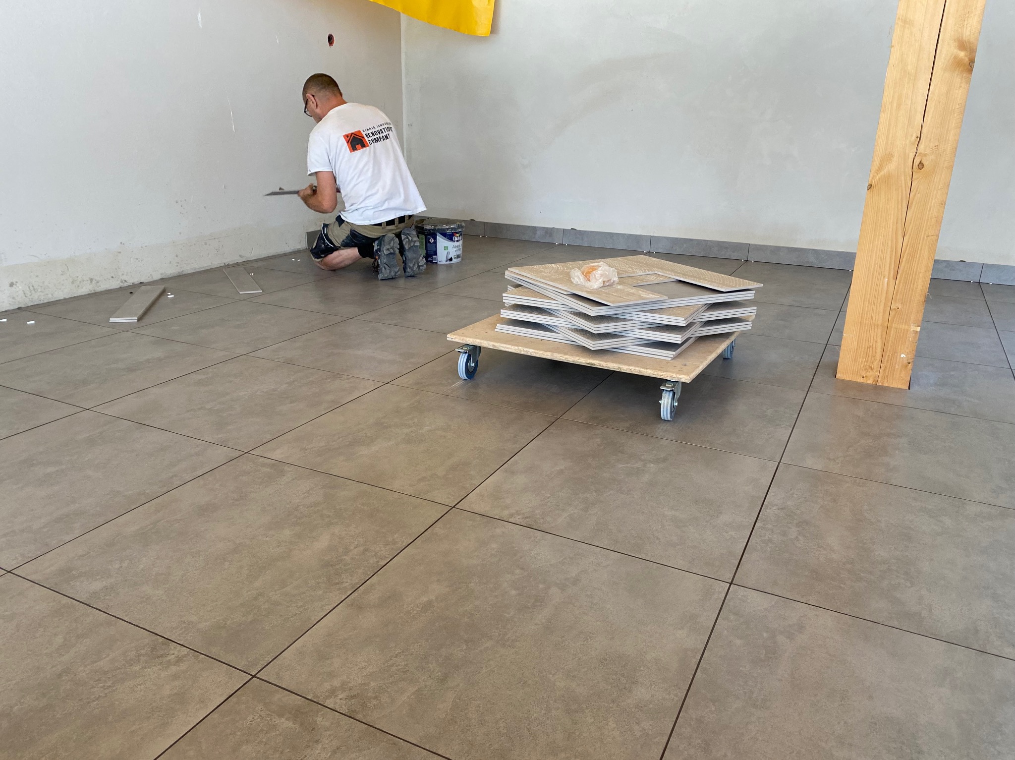 Pracownik w koszulce z logo firmy układa szare płytki podłogowe w pomieszczeniu, na podłodze leżą płytki na wózku na kółkach, widoczne narzędzia i puszki z farbą.