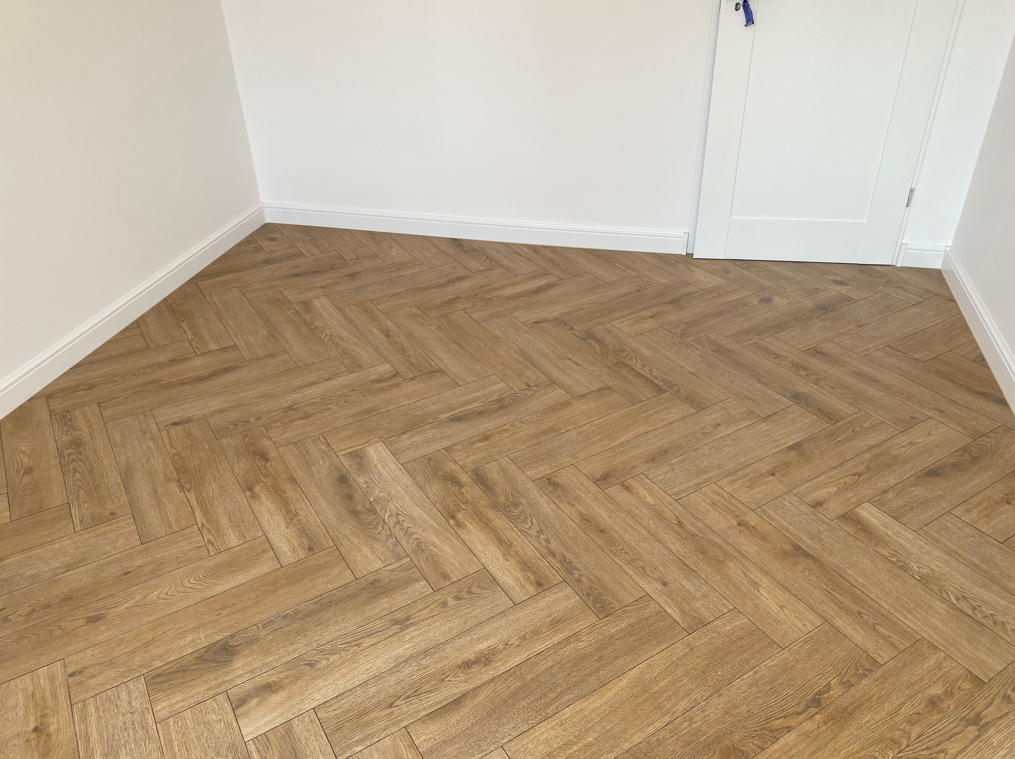 Ułożona pod kątem 45 stopni parkietowa podłoga w odcieniach brązu, z widoczną fakturą drewna, w jasnym pomieszczeniu z białymi ścianami i listwami przypodłogowymi, oraz białe drzwi z niebieską...