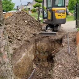 Żółta minikoparka Volvo EC18D przy głębokim wykopie w ziemi, z hałdą ziemi w tle i drzewem po lewej, na częściowo utwardzonym terenie.