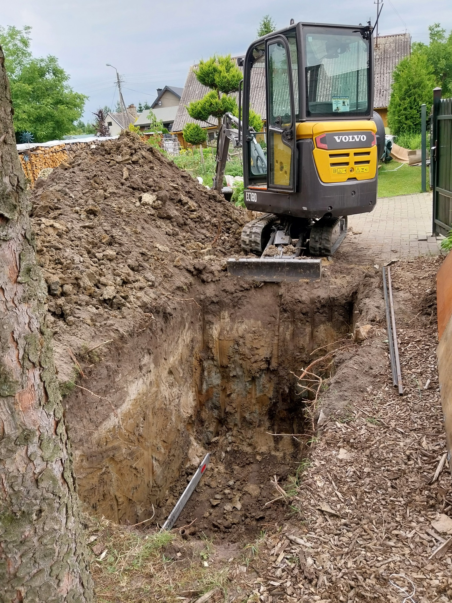 Żółta minikoparka Volvo EC18D przy głębokim wykopie w ziemi, z hałdą ziemi w tle i drzewem po lewej, na częściowo utwardzonym terenie.