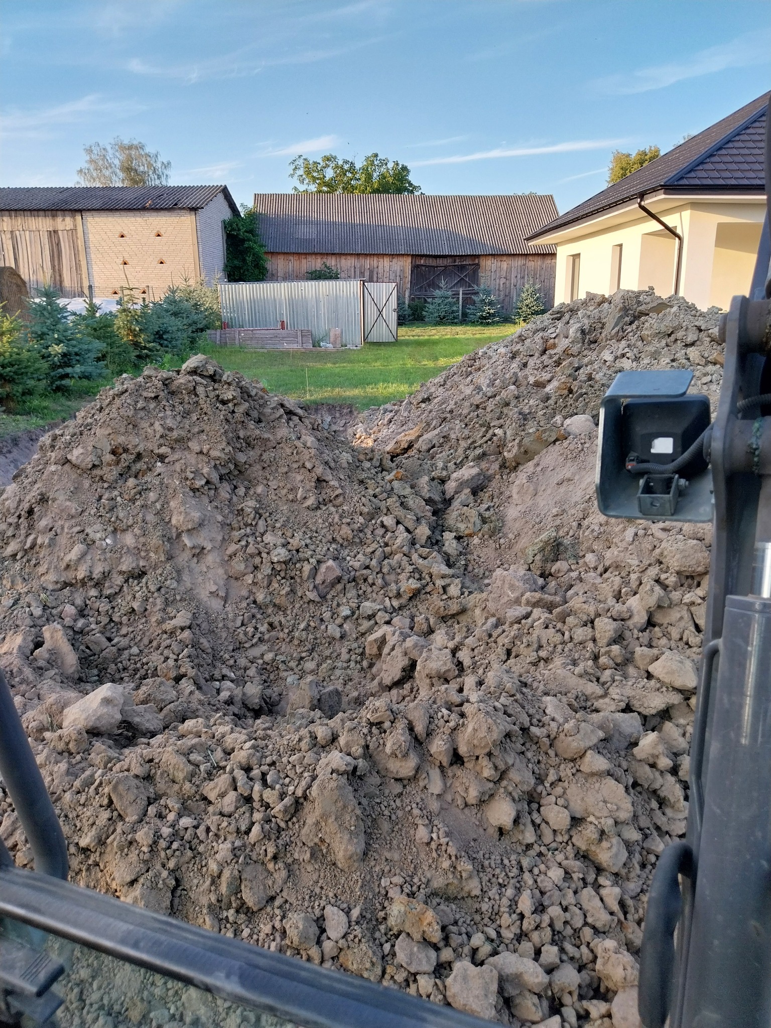 Stos ziemi i kamieni usypany obok wykopu, widoczny fragment maszyny budowlanej, w tle budynki gospodarcze i dom jednorodzinny.