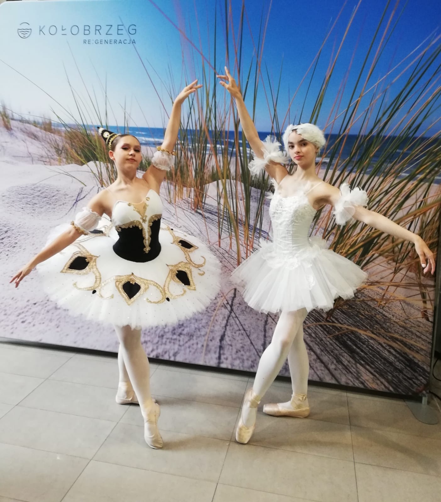 Dwie młode baletnice w kostiumach, pozujące na tle grafiki plaży z napisem 'Kołobrzeg Re:Generacja'.