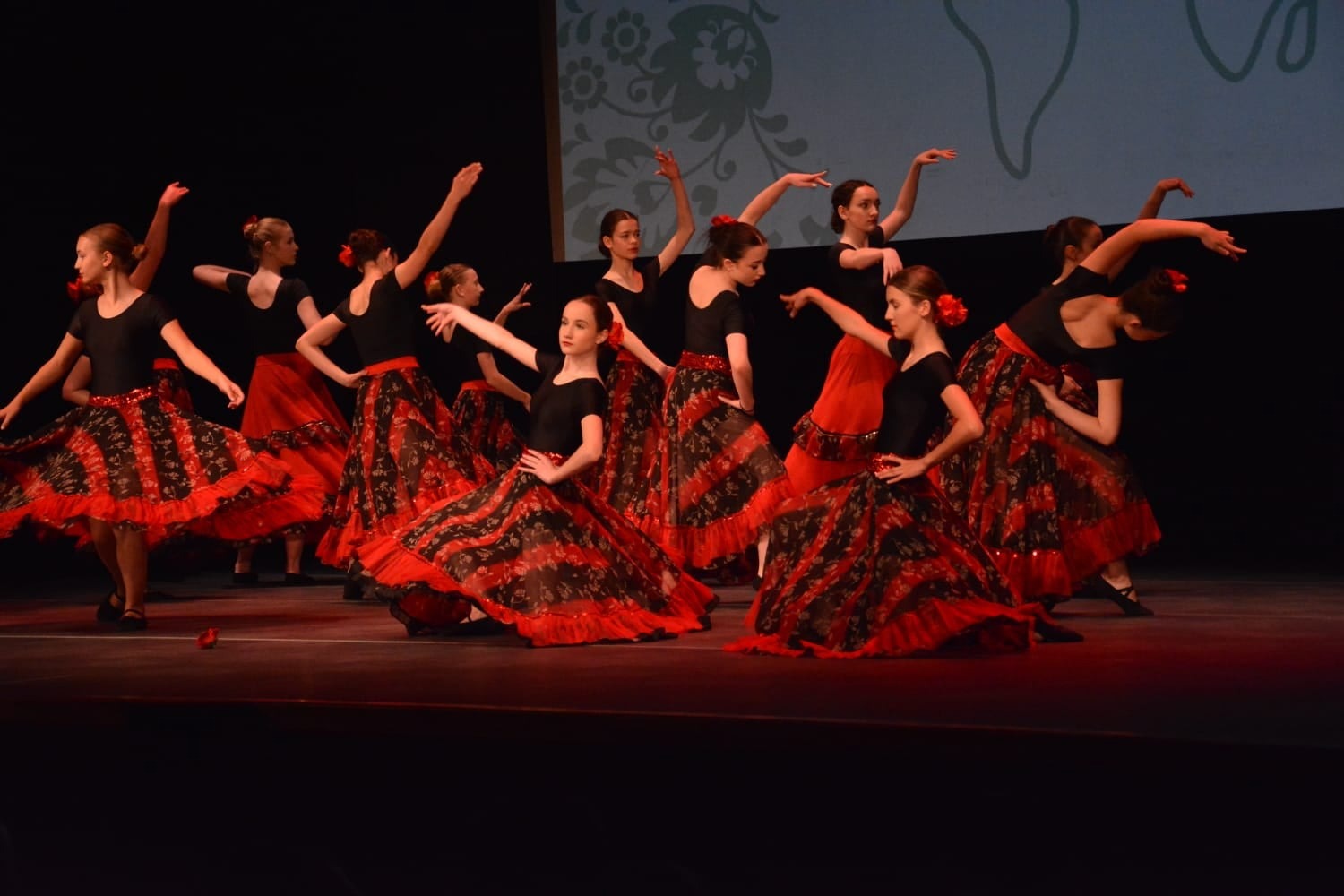 Grupa młodych tancerek w czarno-czerwonych strojach prezentuje układ taneczny na scenie z ciemnym tłem i subtelnym oświetleniem.