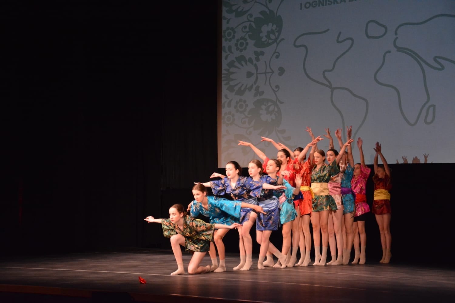 Grupa tancerek w kimonach prezentuje układ na scenie z projekcją mapy świata w tle.