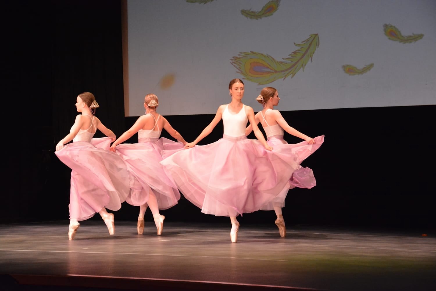 Cztery baletnice w białych strojach i różowych tiulowych spódnicach prezentują zsynchronizowany układ taneczny na scenie z projekcją piór w tle.