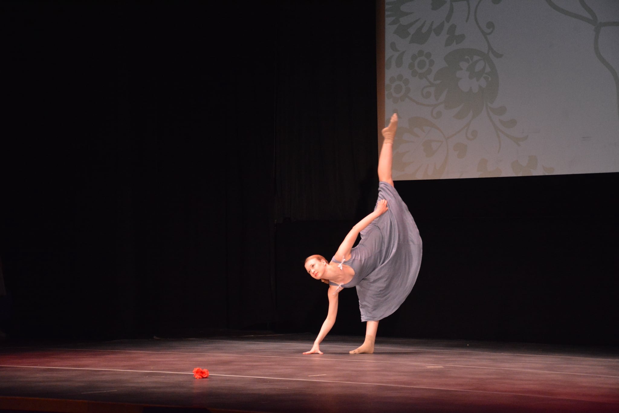Tancerka w szarej sukience wykonuje szpagat na scenie, obok leży czerwony kwiat.