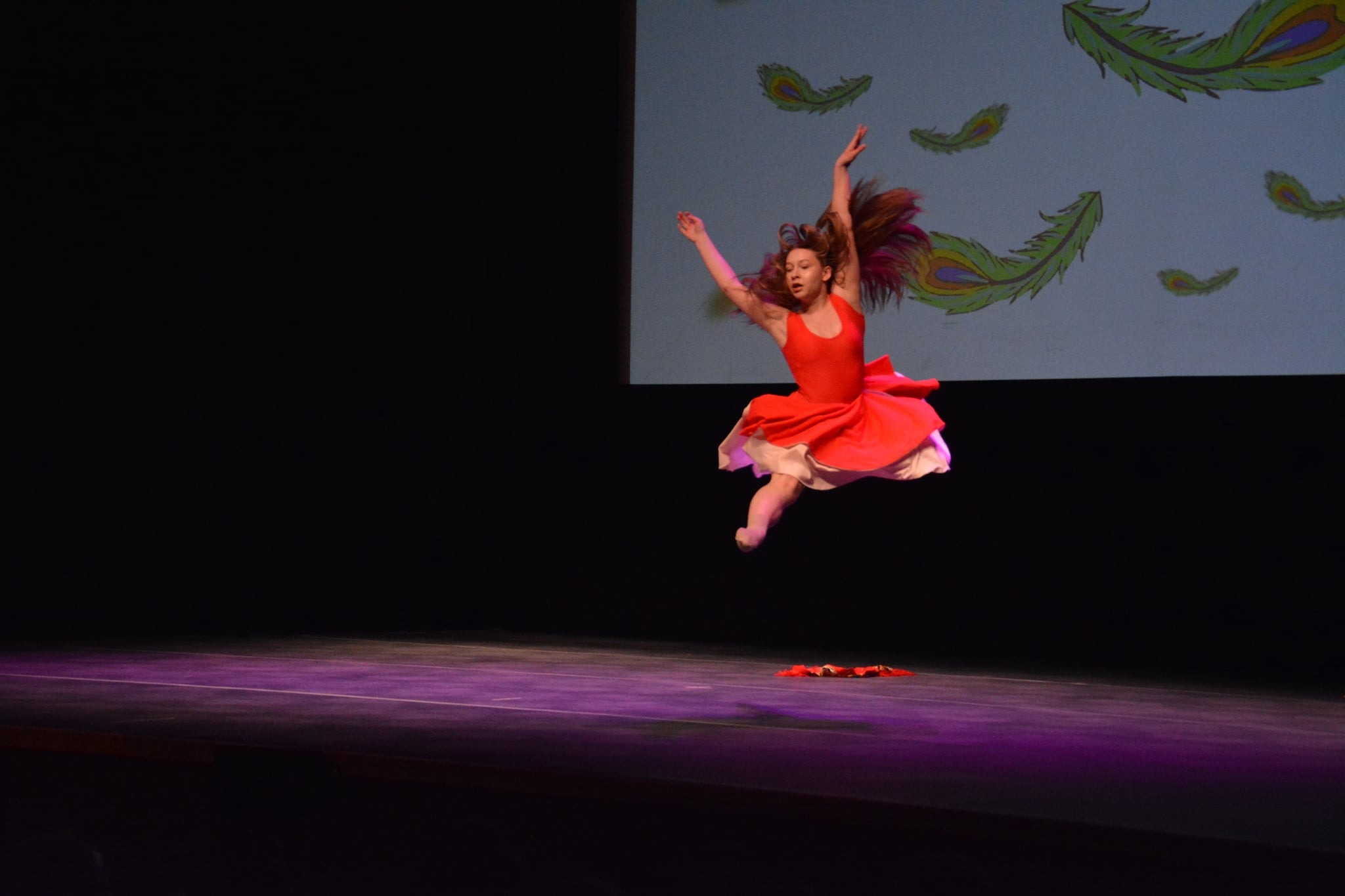 Tancerka w czerwonej sukience w dynamicznym skoku na scenie z projekcją piór na tle.