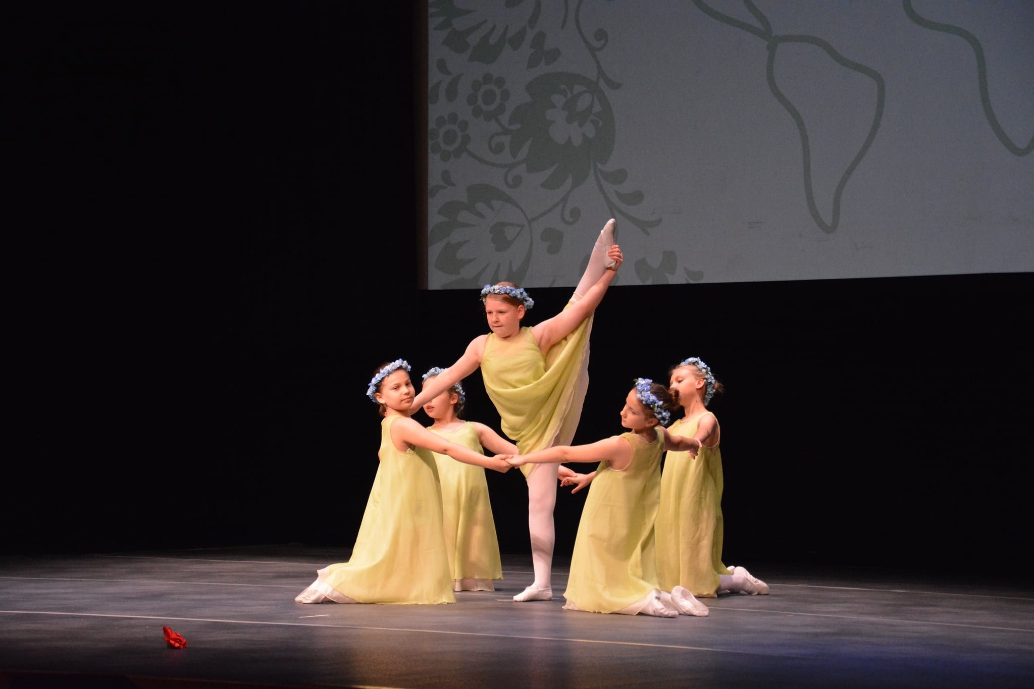 Grupa młodych dziewcząt w jasnożółtych sukienkach i kwiatowych wiankach prezentuje układ taneczny na scenie z ciemnym tłem i wyświetlanym wzorem.