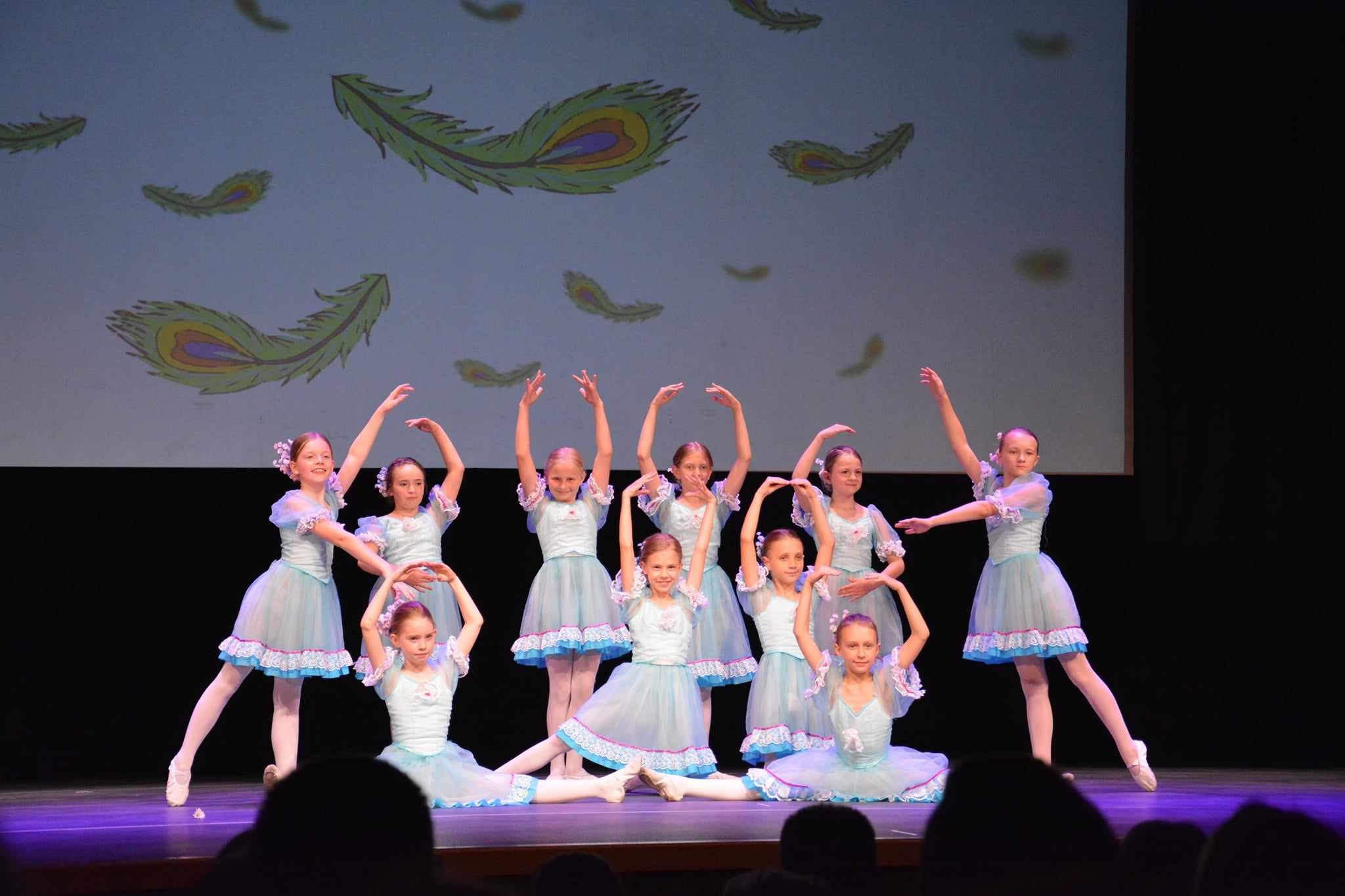 Grupa młodych tancerek baletowych w błękitnych strojach prezentuje układ taneczny na scenie z projekcją pawich piór w tle.