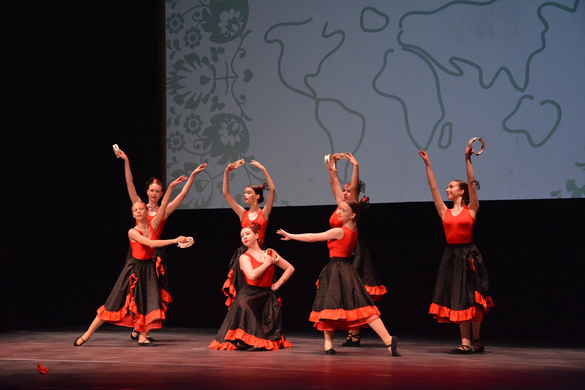 Grupa młodych tancerek w czerwono-czarnych strojach prezentuje układ taneczny z tamburynami na scenie z projekcją mapy świata w tle.