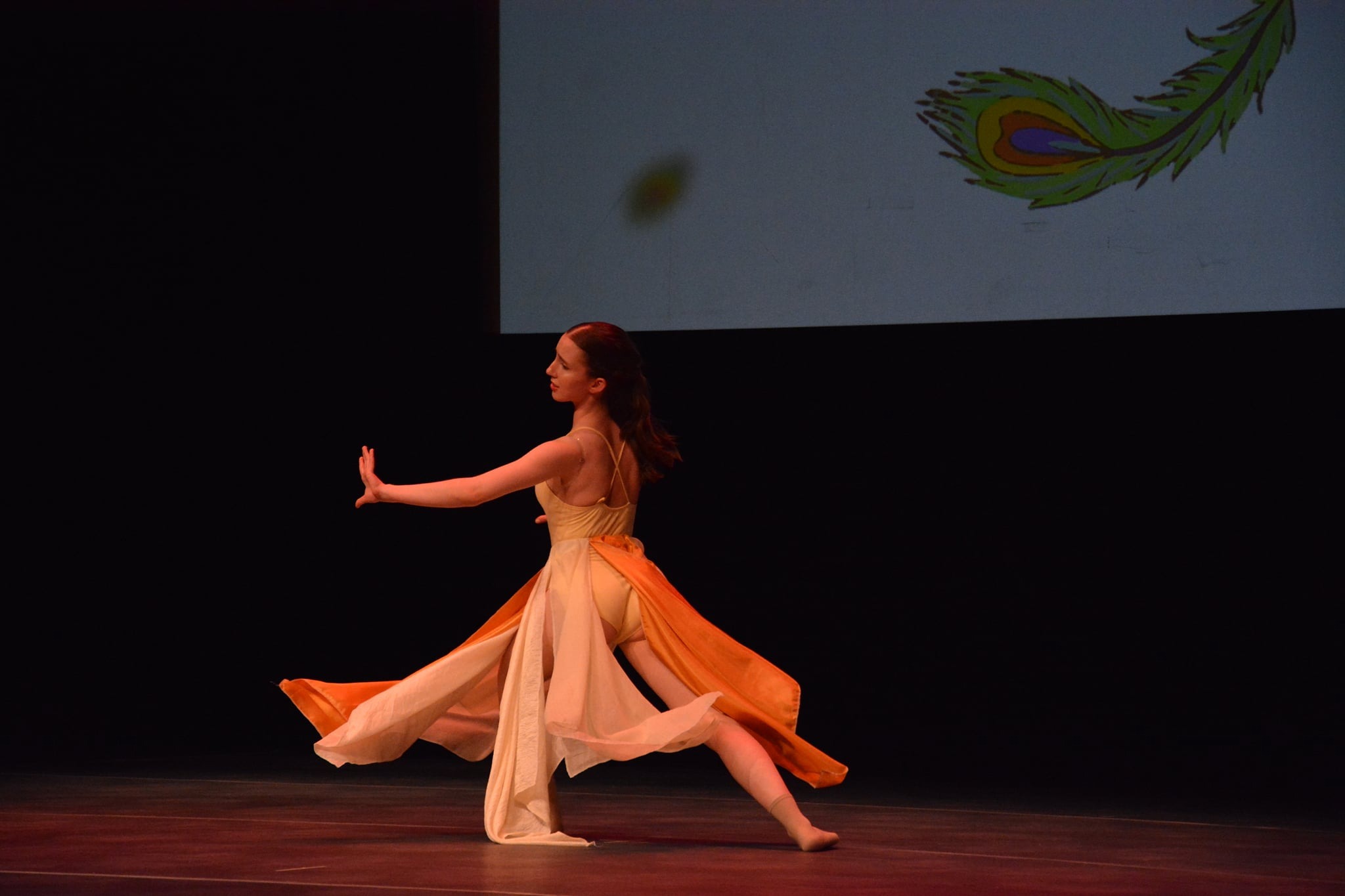 Tancerka w zwiewnej sukience wykonuje dynamiczny ruch na scenie, w tle wyświetlony rysunek pawiego pióra.