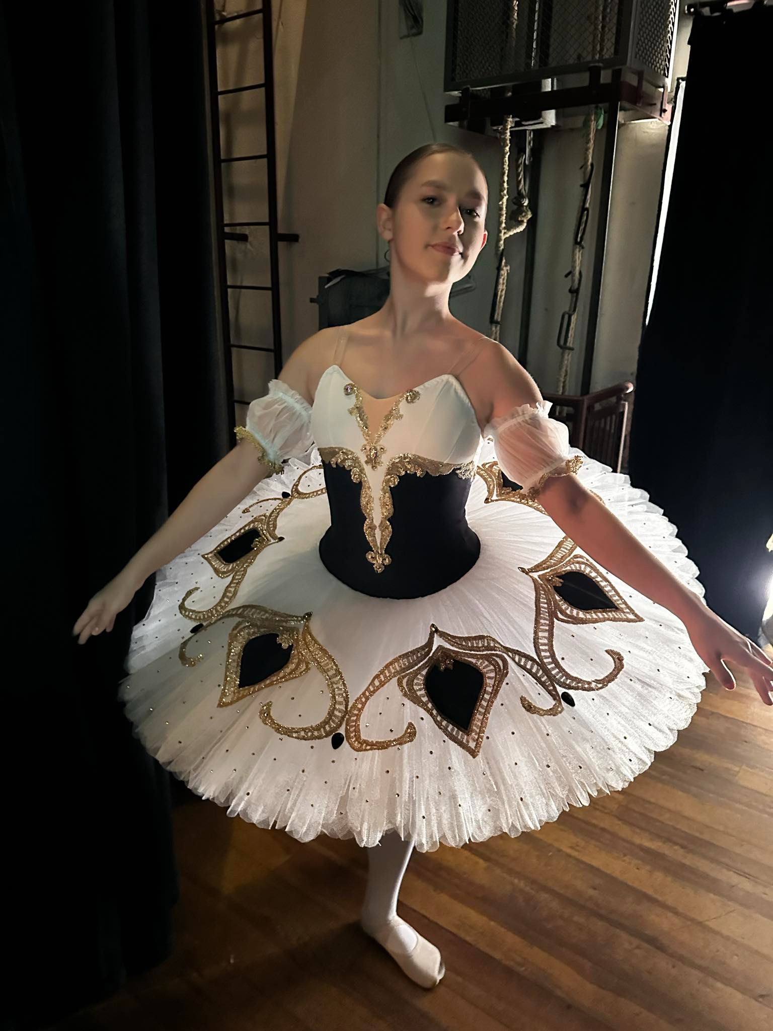 Młoda balerina w kostiumie z tiulem, zdobionym złotymi aplikacjami, pozuje na drewnianej podłodze w teatralnej garderobie z widocznymi rekwizytami.