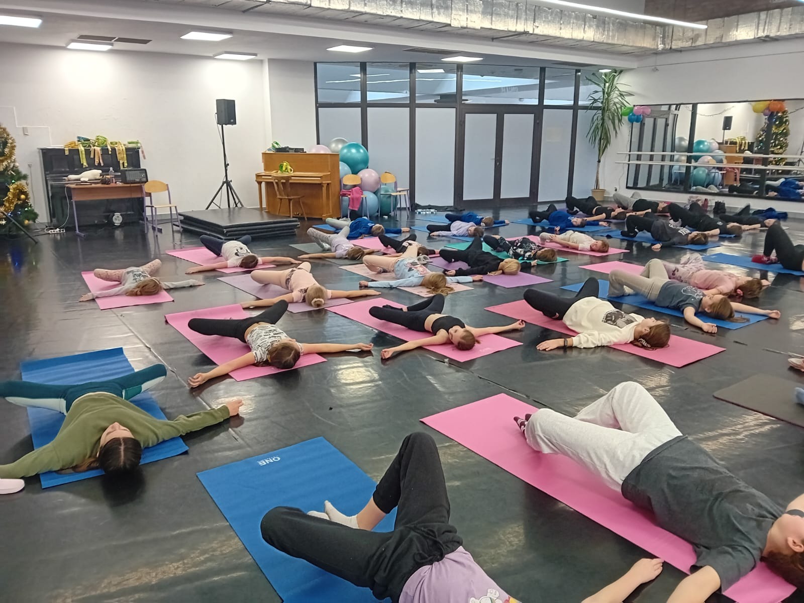 Grupa osób ćwiczy rozciąganie na matach w sali tanecznej; w tle pianino, dekoracje świąteczne i lustra.