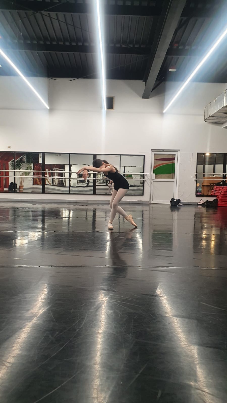 Baletnica w czarnym stroju ćwiczy w sali z lustrami, odbijającymi rośliny, na błyszczącej, czarnej podłodze, oświetlonej przez długie, liniowe lampy sufitowe.