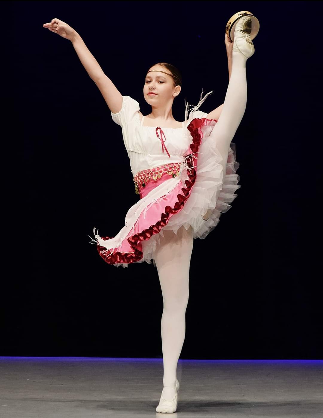 Młoda baletnica w białym stroju z różowymi akcentami, wykonująca wysokie podniesienie nogi na scenie z tamburynem na stopie.