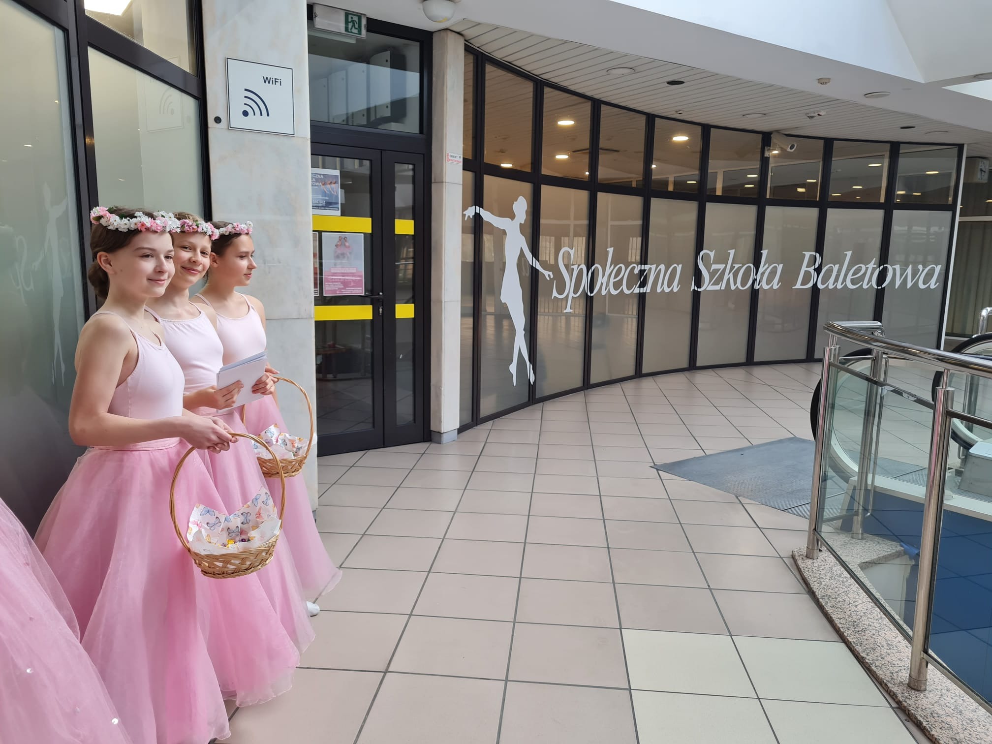 Trzy baletnice w różowych strojach i kwiatowych wiankach, trzymające koszyki, stojące przed budynkiem z napisem 'Społeczna Szkoła Baletowa'.