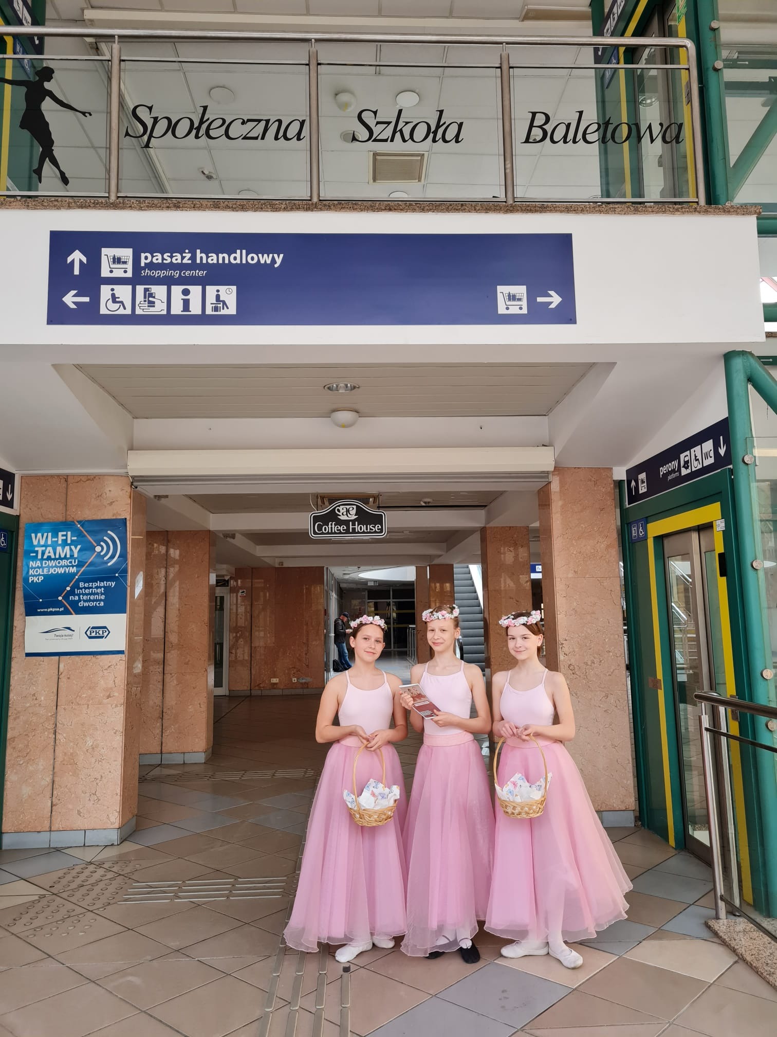 Trzy dziewczynki w różowych baletowych strojach i wiankach na głowach stoją przed wejściem do 'Społecznej Szkoły Baletowej' w przestrzeni dworca kolejowego.