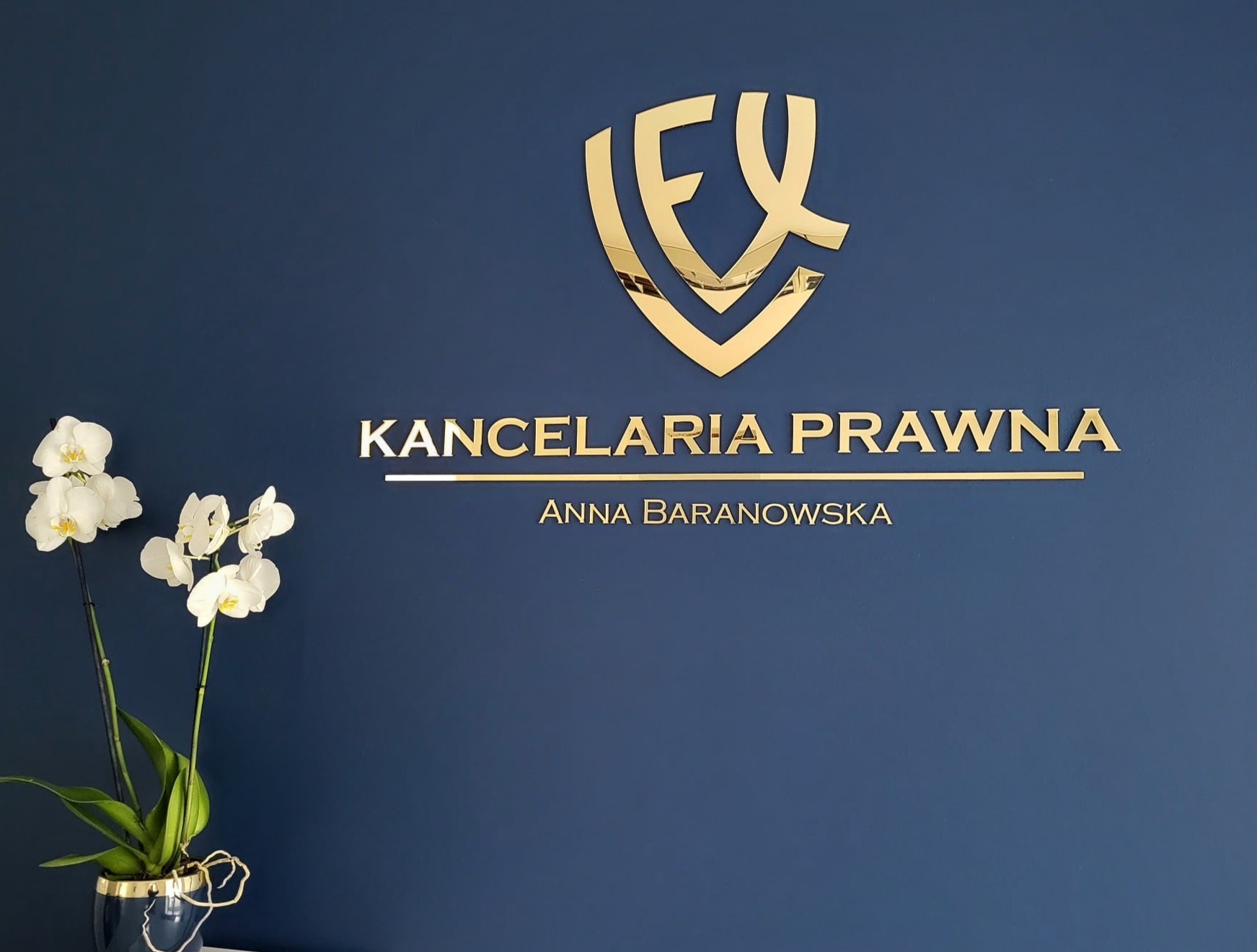 Ściana w kolorze granatowym z złotym logo i napisem 'Kancelaria Prawna Anna Baranowska' oraz białe orchidee w ciemnym wazonie.