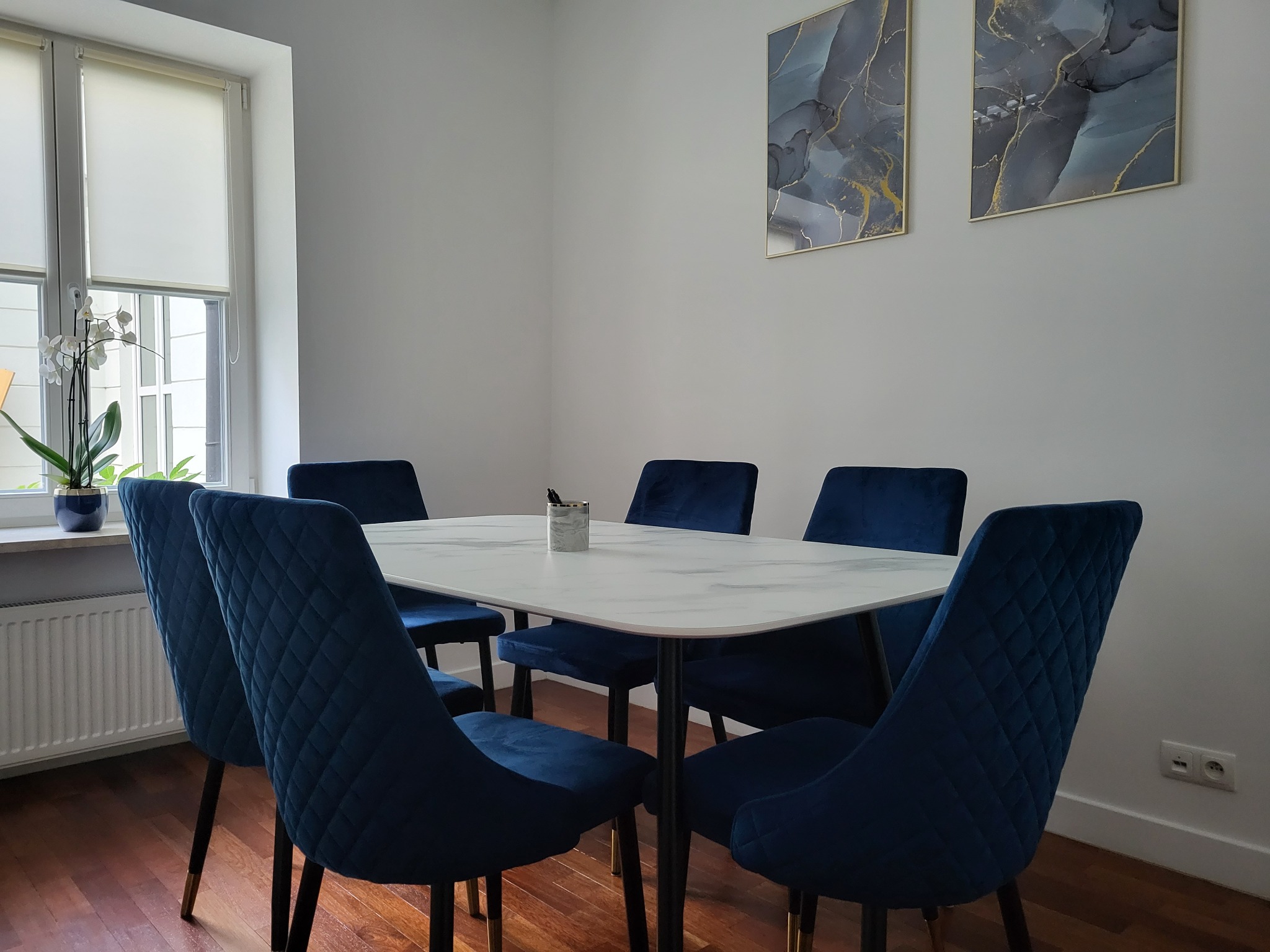 Jadalnia z białym stołem imitującym marmur, sześcioma krzesłami obitymi granatowym welurem z pikowaniem w romby, oraz dwoma abstrakcyjnymi obrazami w szaro-złotej kolorystyce na białej ścianie.