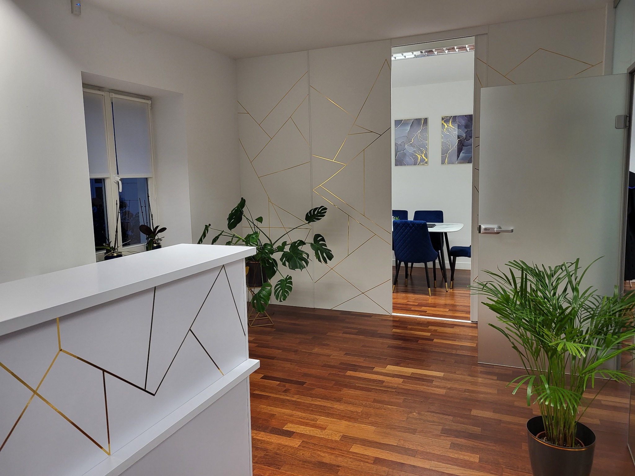 Recepcja biura z białym blatem i złotymi geometrycznymi zdobieniami, widoczne rośliny doniczkowe, parkiet z ciemnego drewna oraz przeszklone drzwi do sali konferencyjnej z niebieskimi krzesłami.
